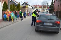 ulica, po chodniku idzie grupa dzieci kolorowo ubranych, policjant zatrzymuje samochody i wręcza ulotki kierowcom