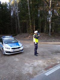 mężczyzna policjant przy użyciu urządzenia mierzy prędkość dla pojazdów, obok stoi policyjny samochód