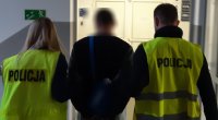 korytarz, kobieta i mężczyzna ubrani w kamizelki odblaskowe z napisem policja prowadzą mężczyznę w kajdankach